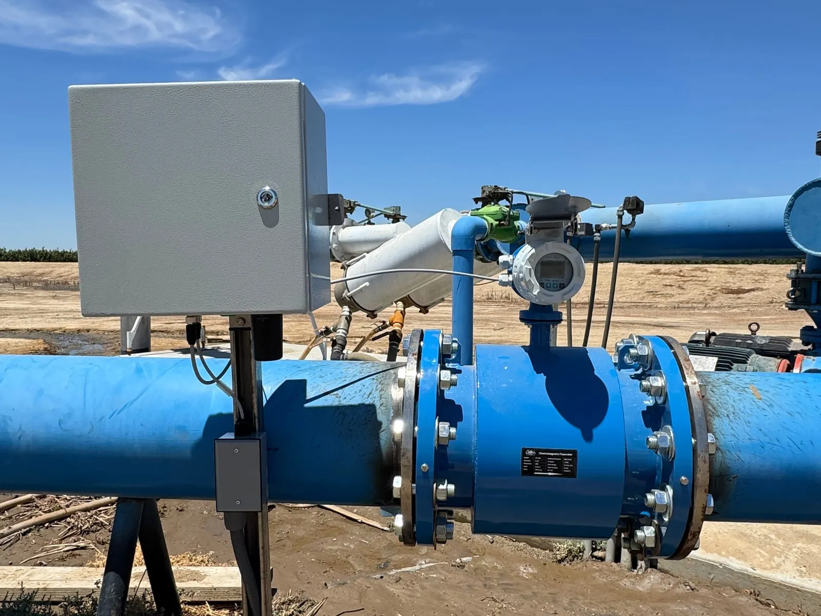 An electromagnetic flow meter on a blue irrigation main beside a telemetry enclosure