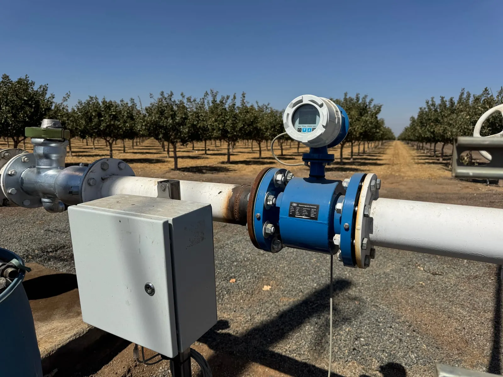 An electromagnetic flow meter with a digital display on a white irrigation main, a telemetry enclosure beside it and orchard rows behind