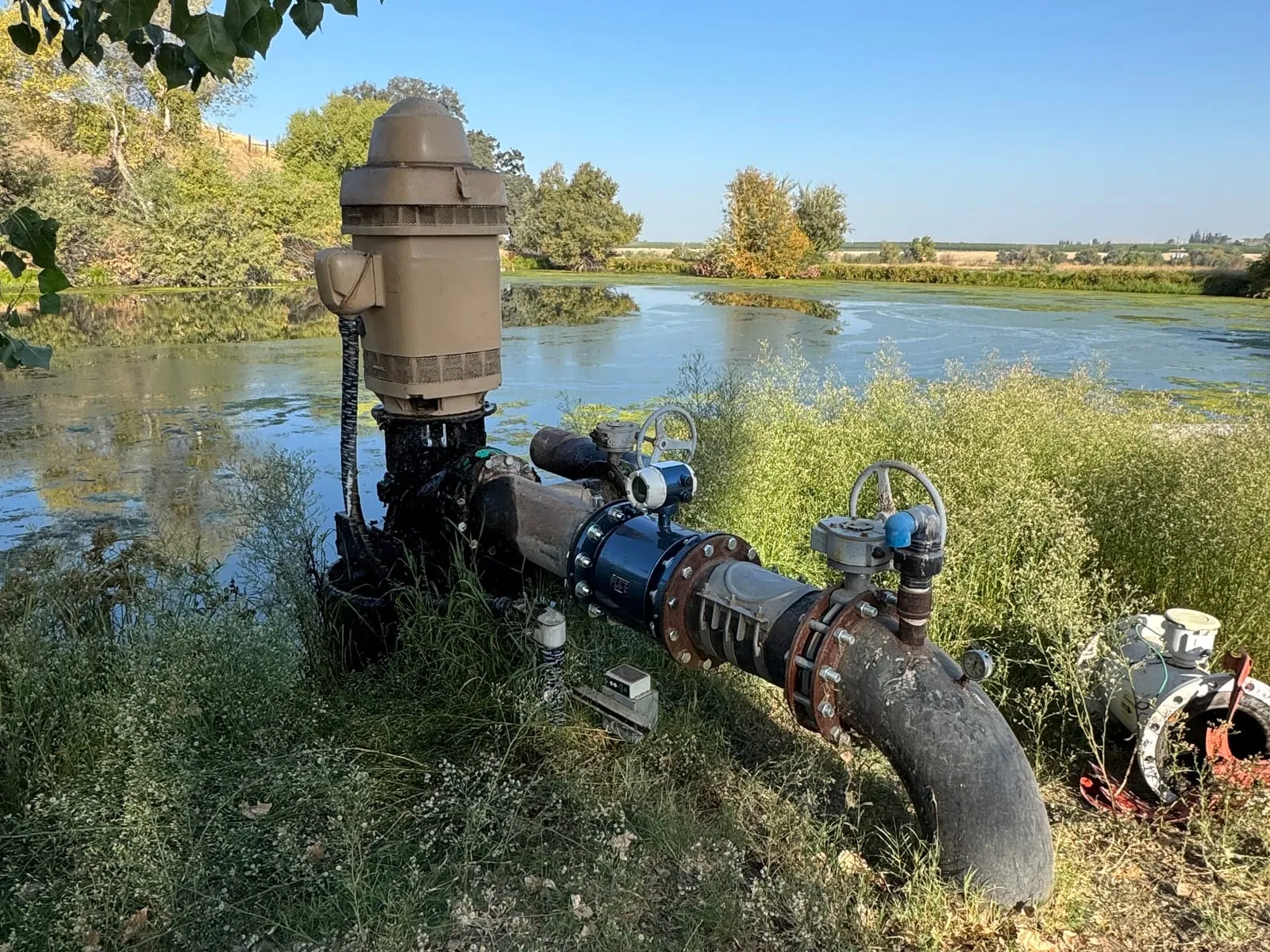 A well pump and discharge line fitted with a magnetic flow meter at the edge of an irrigation pond
